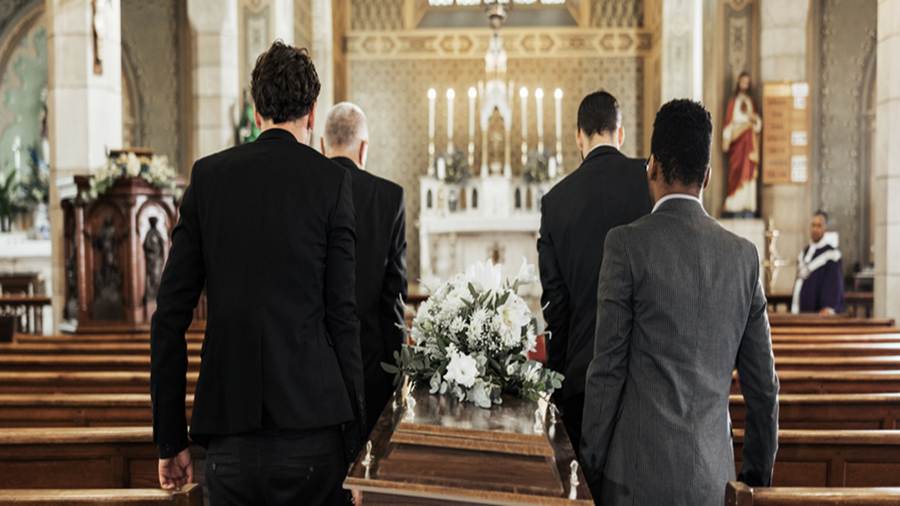 Pallbearers carry a casket inside a church. From behind, the group of men carry the white flower covered coffin toward the altar.